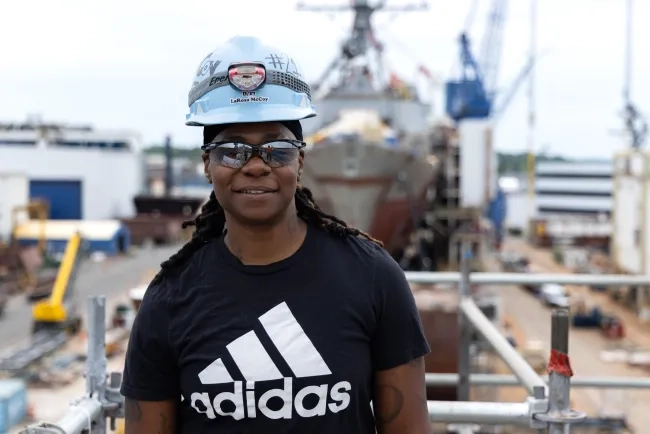 Black female with helmet and headlamp and foreground, with ship and shipyard equipment in background